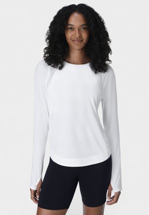 Vrouw met krullend haar, gekleed in een wit shirt met lange mouwen en zwarte fietsbroekjes, staand en glimlachend tegen een effen achtergrond.