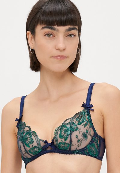 Nainen pitsinen rintaliivi, jossa on vihreä kukkakirjonta läpinäkyvässä verkkokankaassa, laivastonsinisiä hihnoja koristetupsuilla ja kolmionmuotoiset kupit.