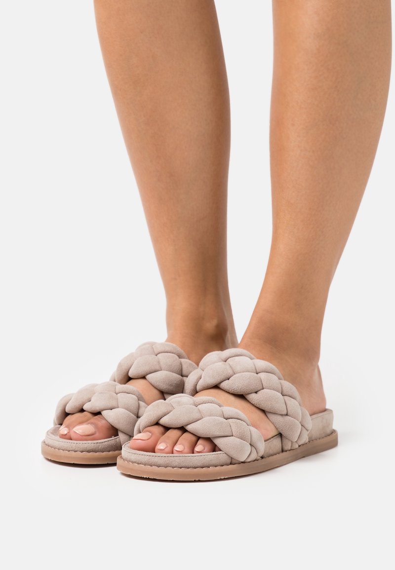 Pieds portant des sandales beige à brides tressées avec semelles plates sur fond blanc.