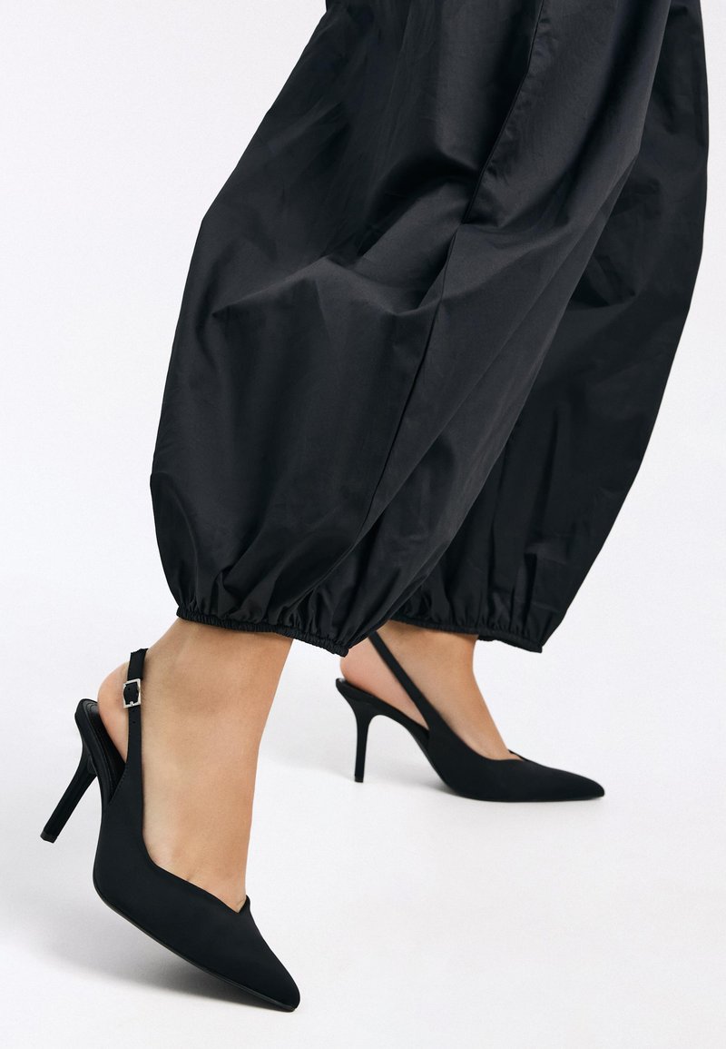 Pantofi cu toc slingback negri cu vârfuri ascuțite, tocuri înalte de tip stiletto și un finisaj mat neted, asortați cu pantaloni negri, elastici, de tip balon.