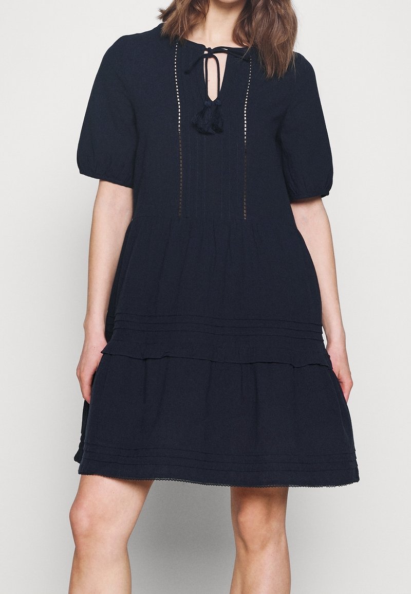 Tmavě modré šaty vyrobené z texturované tkaniny, s volánovým designem, krátkými nabíranými rukávy a výstřihem ve tvaru klíčové dírky s detailem na zavazování pomocí třásní.