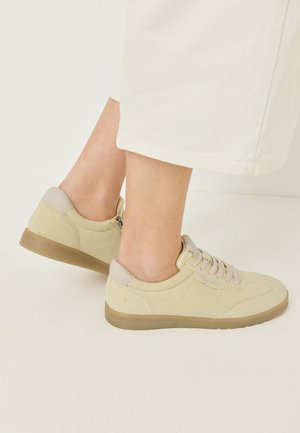 Pies con zapatillas de ante beige con cordones y suelas marrones, combinadas con pantalones cortos color crema sobre un fondo claro.