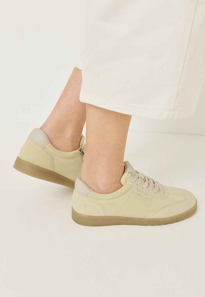 Πόδια με μπεζ σουέτ sneakers με καφέ σόλες, συνδυασμένα με κρεμ cropped παντελόνια σε ανοιχτό φόντο.