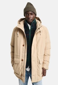 Beżowa parka z kapturem, zapinana na zamek i guziki, z dwiema przednimi kieszeniami i zieloną polarową bluzą zapinaną na zamek pod spodem, noszona z prążkowanym beanie.