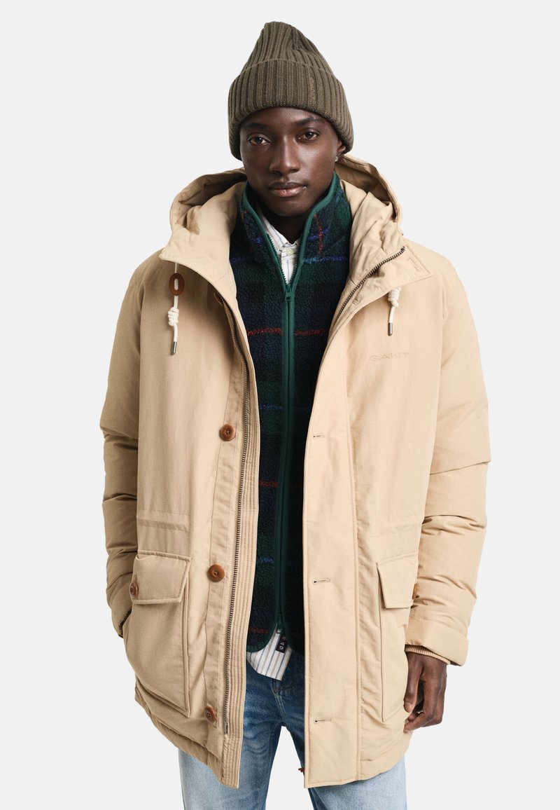 Beżowa parka z kapturem, zapinana na zamek i guziki, z dwiema przednimi kieszeniami i zieloną polarową bluzą zapinaną na zamek pod spodem, noszona z prążkowanym beanie.