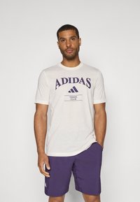 Kremfarget T-skjorte med kort erm og lilla "ADIDAS TENNIS CLUB" trykk, kombinert med lilla shorts. Laget av bomull, løs passform, rund hals.