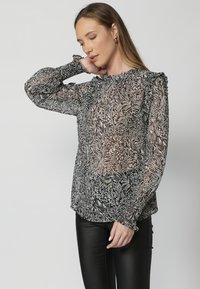 Fekete-fehér virágmintás áttetsző blúz fodros mandzsettával, kerek nyakkivágással és laza fazonnal. Fekete, slim fit nadrággal párosítva.