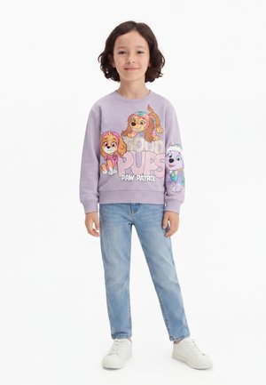Barn stående iført en lilla Paw Patrol sweatshirt med hundefigurer, lyseblå jeans og hvide sneakers på hvid baggrund.