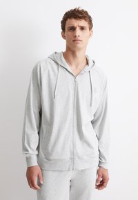 Ljusgrå zip-up huvtröja i mjukt tyg, med en huva, kängurufickor och ribbade manschetter, samt en liten logotyp på bröstet.