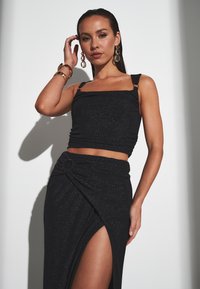 Sort glitrende crop top med justerbare stropper og tætsiddende design, parret med en knyttet sort nederdel med en sidelæg.