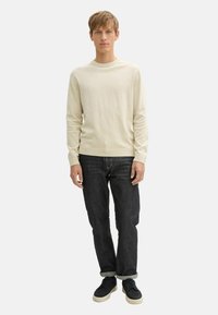 Maglione beige chiaro realizzato in materiale knit con scollo rotondo, abbinato a jeans in denim scuro e sneaker nere.