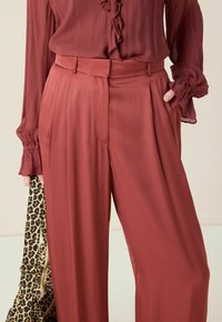 Vrouw in roestkleurige geplooide broek en bijpassende blouse met ruffles, die met één hand een tas met luipaardprint vasthoudt terwijl ze met de andere hand in haar zak zit.