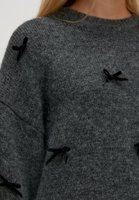 Grauer gestrickter Pullover mit strukturierter Oberfläche, verziert mit kleinen schwarzen Samtschleifen als Akzente am Körper und an den Schultern. Lässiger Schnitt.