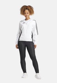 Witte zip-up jas met zwarte zijstrepen, gedragen over zwarte leggings. Het model staat in een neutrale houding tegen een plain achtergrond.