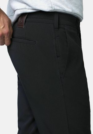 Person trägt schwarze Hosen mit seitlichen und hinteren Einschubtaschen, greift in die hintere Tasche, grau shirt teilweise sichtbar.