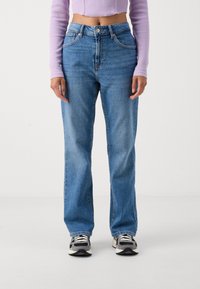 Jeans de mezclilla de color azul claro con un ajuste relajado, cintura alta, diseño de cinco bolsillos y sutiles efectos de desvanecimiento, combinados con zapatillas blancas y negras.