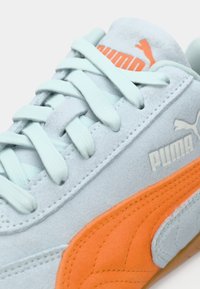 Sapatilhas de camurça azul claro com detalhes em laranja, atacadores planos e sola de borracha com um logo distintivo da Puma na lateral.