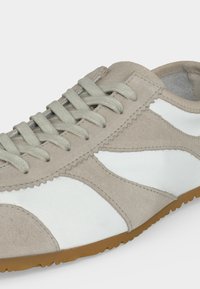 Ανοιχτό μπεζ και λευκό αθλητικό παπούτσι με σουέντ λεπτομέρειες, μπεζ κορδόνια και καφέ λαστιχένια σόλα, εμφανιζόμενο από πλευρική γωνία.