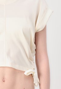 Top corto beige chiaro con maniche corte arrotolate, lati arricciati e lacci regolabili. Texture liscia con una leggera lucentezza.