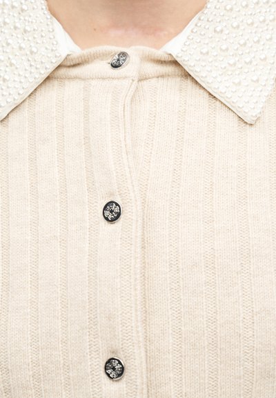 Cardigan en maille côtelée crème avec des boutons floraux noirs et un col pointu blanc orné de petites perles.