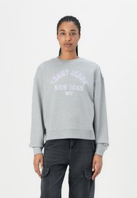 Felpa oversize grigia chiara con collo a giro e polsini a costine, con la scritta "TOMMY JEANS NEW YORK CITY" ricamata in viola.