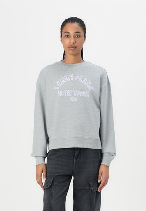 Gaiši pelēks, pārāk liels džemperis ar apaļu kakla izgriezumu un ribinātām aprocēm, ar violetā krāsā izšūtu uzrakstu "TOMMY JEANS NEW YORK CITY".