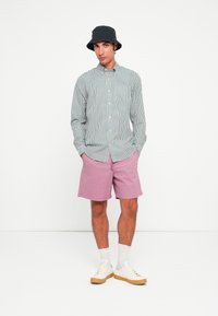 Stribet skjorte i grønt og hvidt, pink shorts, grå sokker, hvide sneakers med gummisåler og en sort bøttehat.