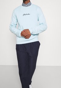 Sudadera azul clara con "Ámsterdam" bordado en negro. Usada sobre una camisa de cuadros. Combinada con pantalones azul marino oscuros y zapatillas blancas.