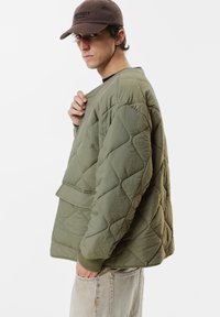 Quilted olivengrøn jakke med fremtrædende diamantmønster, ribbede manchetter og en frontlomme. Stof virker tekstureret og let.