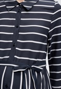 Φόρεμα σε ναυτικό μπλε με λευκές οριζόντιες και κάθετες ρίγες, εμπρόσθιο κουμπωτό, γιακά στον λαιμό, μακριά μανίκια και ζώνη στη μέση που δένει.