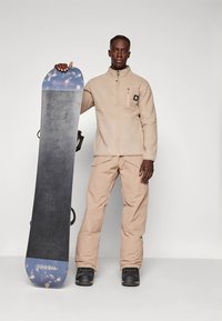 Beżowa polarowa kurtka na zamek błyskawiczny z kieszenią, zestawiona z beżowymi spodniami narciarskimi. Czarna deska snowboardowa z niebieskimi akcentami trzymana w pozycji stojącej.