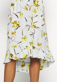 Blumenkleid mit weißem Grundstoff, verziert mit gelben und braunen Blumenmustern. Der Saum hat eine geraffte Kante, die eine geschichtete Textur erzeugt.