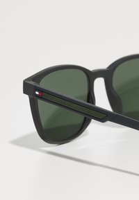 Ochelari de soare negri cu lentile verzi, având o finisare mată, un design unghiular și un accent texturat verde pe brad, alături de un logo roșu și albastru.