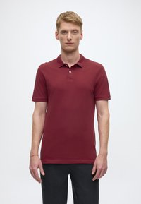 Maroon polo shirt with a three-button placket, short sleeves, and a smooth texture, paired with dark gray trousers.