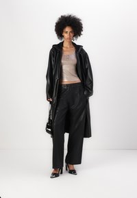 Palton lung din piele synthetică neagră peste un top bej lucios, asortat cu pantaloni negri lejeri și tocuri negre cu accente argintii. Include o poșetă neagră.