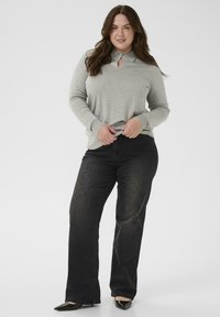Vrouw met lang bruin haar, gekleed in een grijs langarmshirt met een sleutelgatdetail, zwarte wijde jeans en zwarte puntige schoenen, staande.