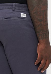 Marineblaue Hose mit glatter Textur, mit einer Gesäßtasche und einem Etikett auf dem Bund. Hand an der Seite sichtbar.
