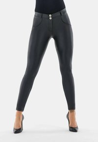 Femme portant un pantalon skinny en simili cuir noir et des talons hauts noirs, debout les jambes écartées sur fond blanc.