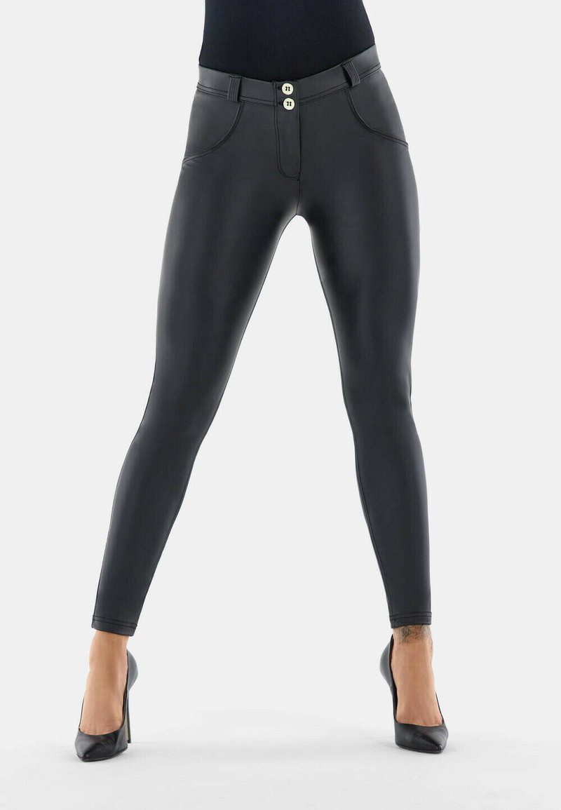 Femme portant un pantalon skinny en simili cuir noir et des talons hauts noirs, debout les jambes écartées sur fond blanc.