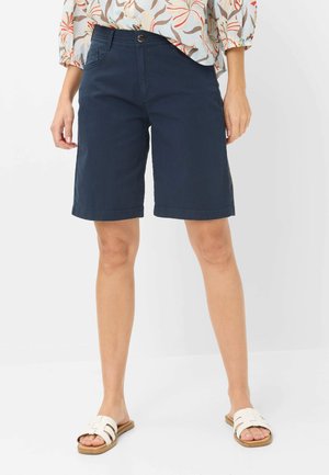 Kvinde iført marineblå knælange shorts, blomstret bluse og hvide slip-on sandaler, stående mod en ensfarvet baggrund.