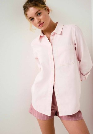 Young woman with blonde hair wearing a light pink button-up shirt and mauve striped shorts standing against a plain white wall.