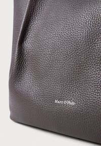 Sac à main en cuir marron texturé présentant le nom de la marque "Marc O'Polo" en blanc, situé dans le coin inférieur droit.