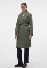 Lange, olijfgroene wikkeljas van zacht materiaal, met een ceintuur in de taille, een reverskraag en twee voorzakken. Gestyled met donkere jeans en laarzen.