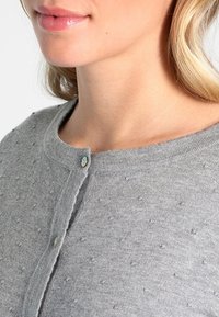 Kvinna klädd i en ljusgråstickad tröja med små upphöjda prickar, knäppt nära kragen, som visar läppar samt del av hals och hår.