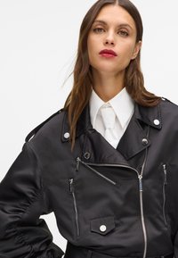 Giacca bomber in raso nero con tasche con cerniera, finiture argento, spalline sulle spalle e vestibilità comoda; indossata sopra una camicia bianca con colletto.