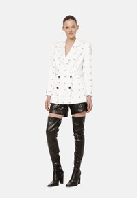 Biała podwójnie zapinana marynarka z czarnymi wzorami, zestawiona z błyszczącymi czarnymi skórzanymi szortami i kozakami do uda, charakteryzująca się eleganckim fasonem.