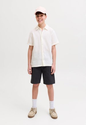 Smilende gutt iført en lys rosa caps, hvit kortermet knappe-skjorte, svarte shorts, hvite sokker og beige sko, stående på en ensfarget bakgrunn.
