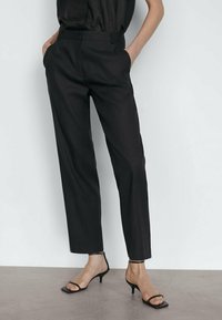 Pantalon noir sur mesure à jambe droite, avec une légère brillance, tissu lisse, poches latérales, accompagné de sandales noires à talons minimalistes.