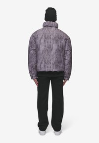 Gewatteerde jas in paars textiel met een hoge kraag. Gecombineerd met zwarte broek en witte sneakers, onder een zwarte beanie.