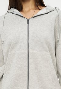 Κοντινό πλάνο ενός ατόμου που φοράει ανοιχτό γκρι φούτερ με φερμουάρ, με μπροστινή τσέπη και κουκούλα με κορδόνι, σε συνδυασμό με ένα κολιέ.